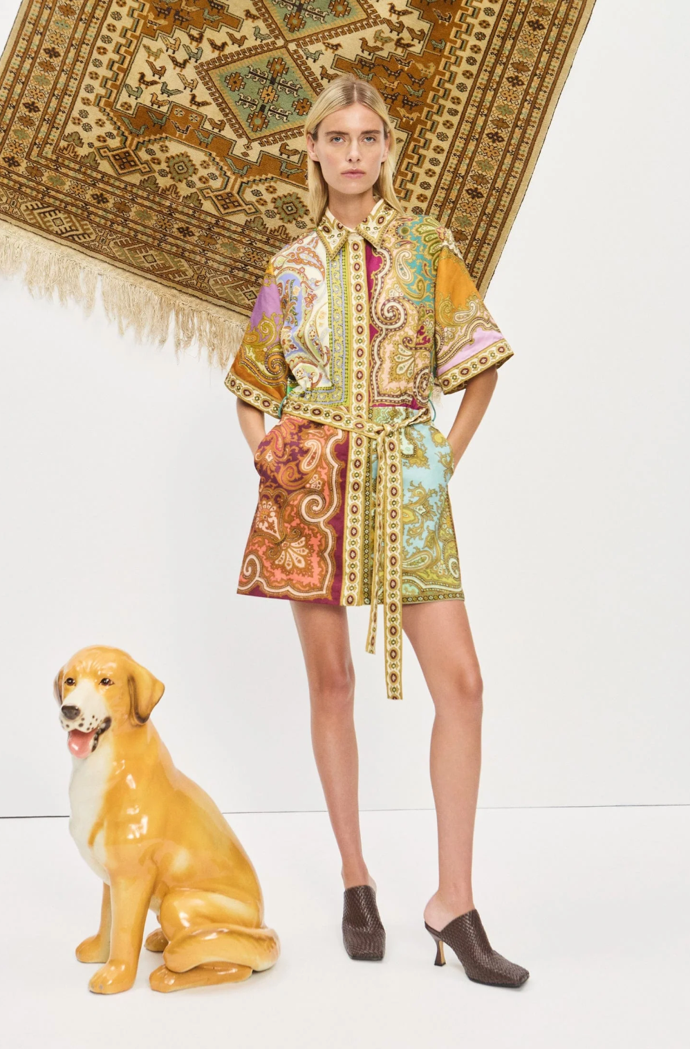 Woman in a patterned dress standing next to a decorative dog statue with a patterned rug in the background.