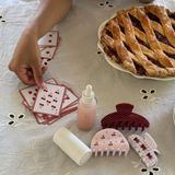 Table with playing cards, a pie, a bottle, and hair accessories on a floral tablecloth.