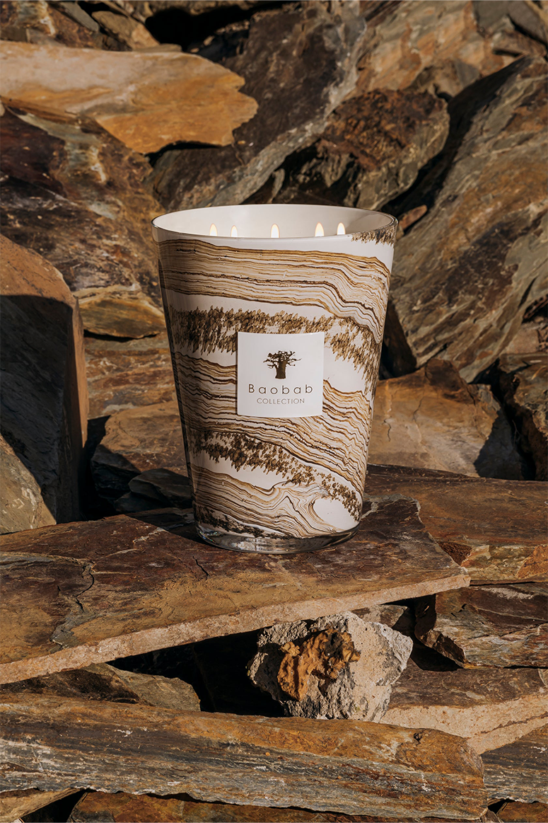 A candle with beige and brown shades in a glass holder, placed on a rocky surface.