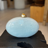 Light blue ceramic clutch with gold clasp on top on a dark surface, blurred indoor background