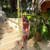 Woman in a terracotta palm print dress standing on a wooden deck with plants and a building in the background