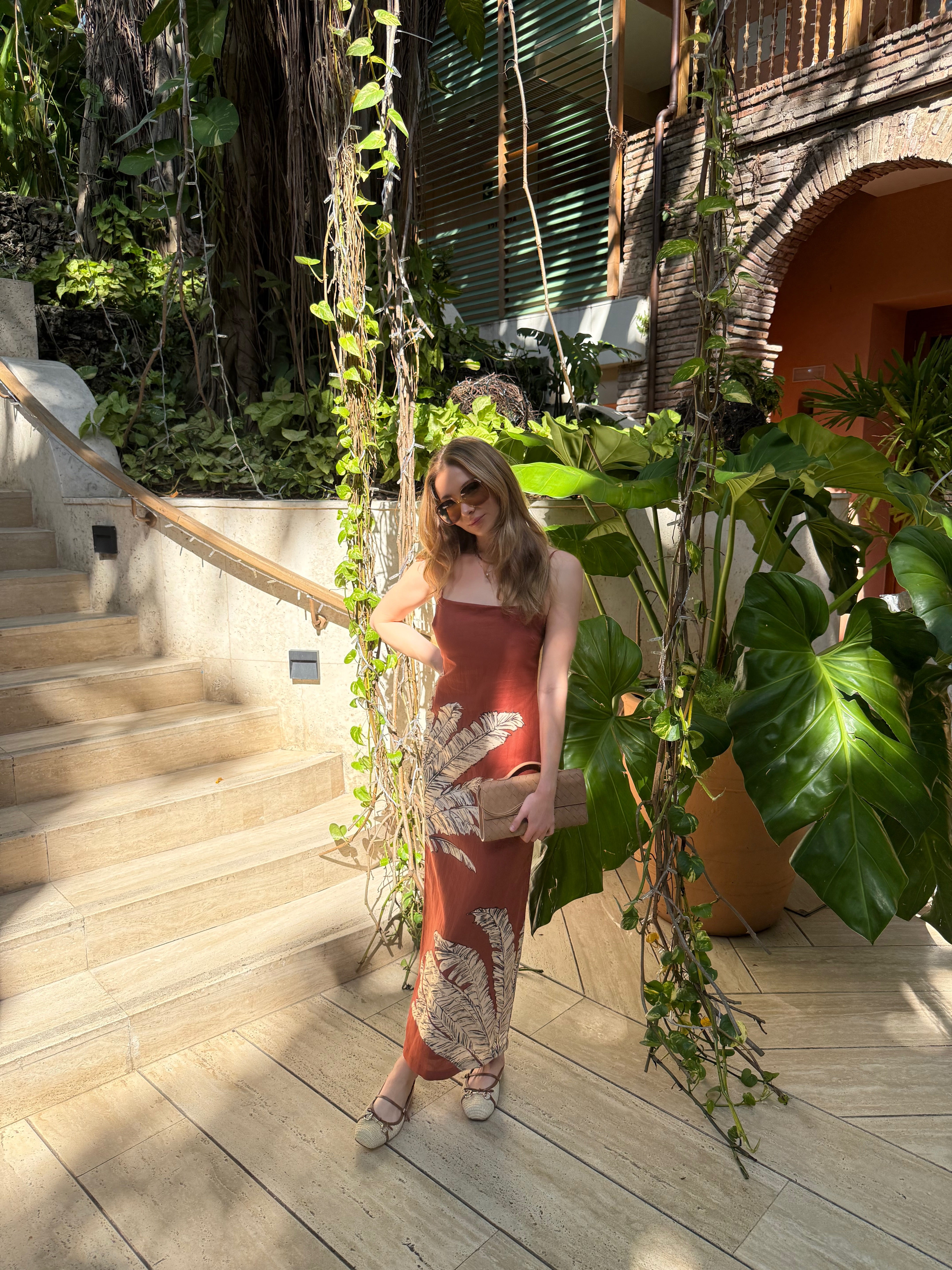 Woman in a terracotta palm print dress standing on a wooden deck with plants and a building in the background