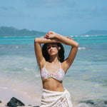 Woman in a floral bikini top and white sarong standing on a beach with clear blue water and sky.