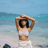 Woman in a floral bikini top and white sarong standing on a beach with clear blue water and sky.