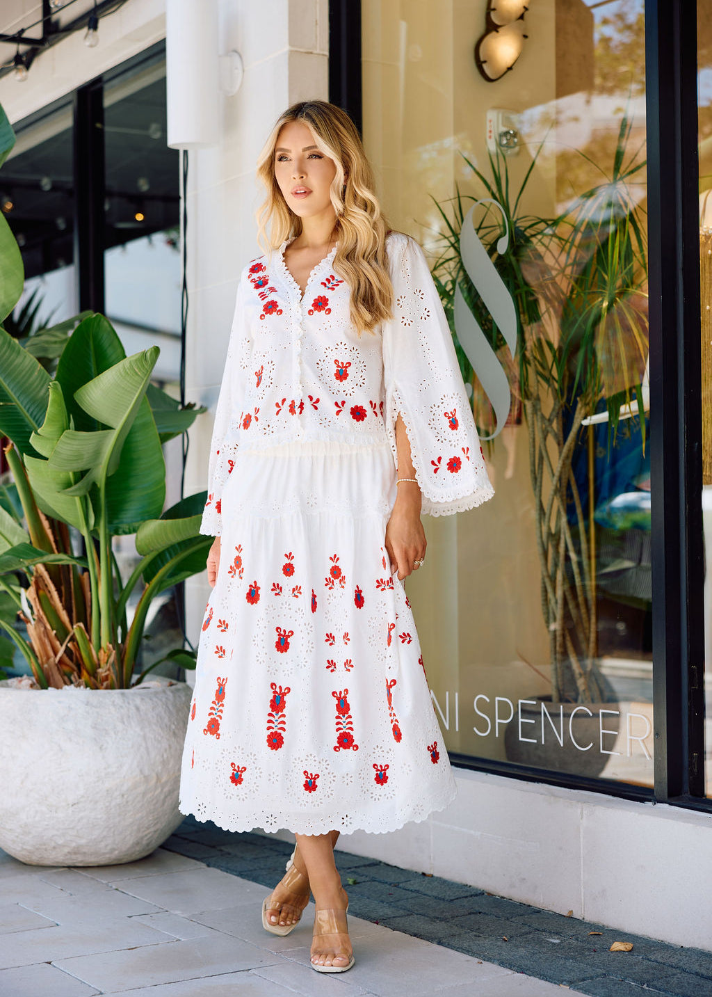 Woman in a white dress with red embroidery standing in front of a store window.
