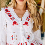 Woman wearing a white blouse with red floral embroidery in an indoor setting.