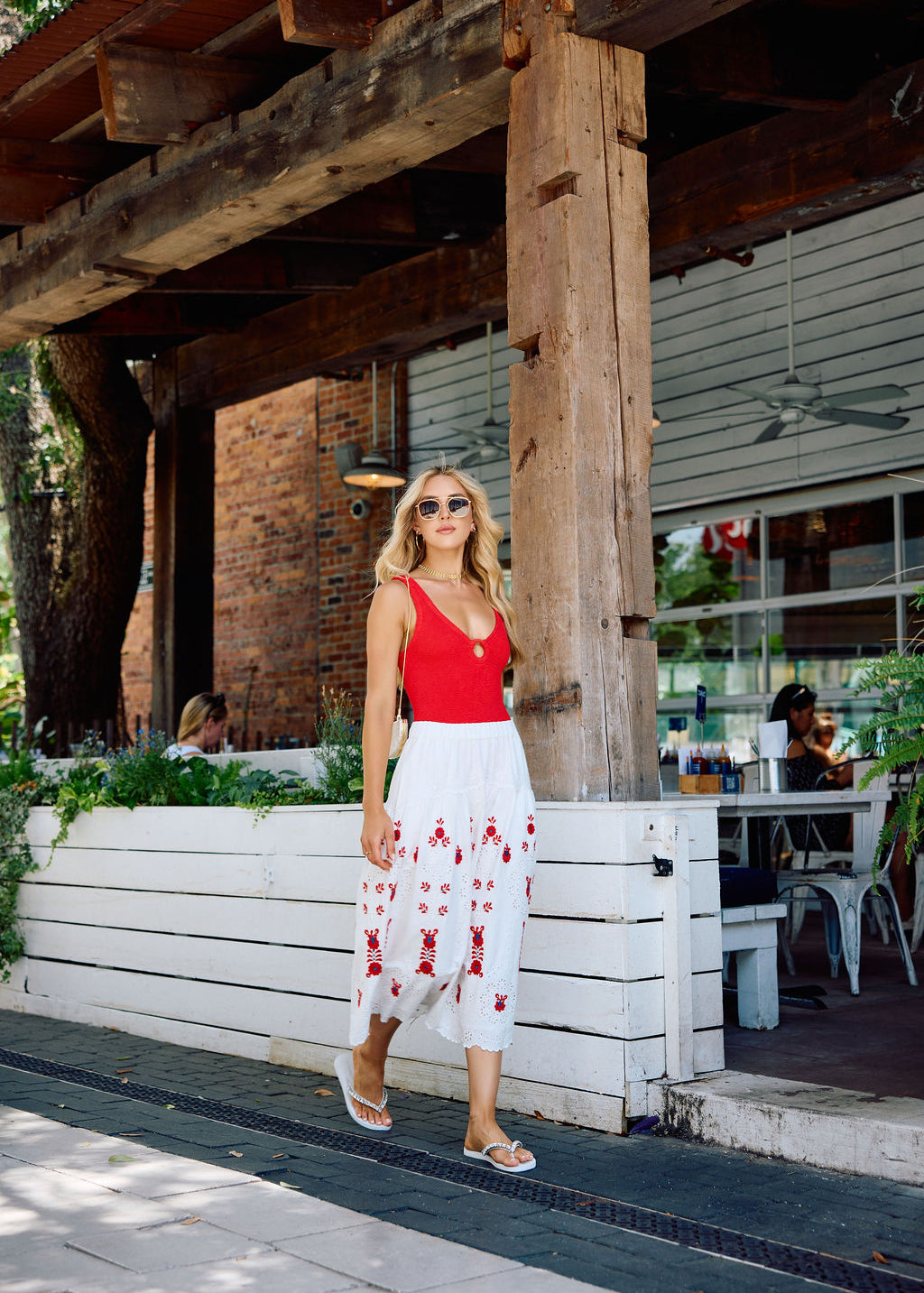 Woman in a red top and white skirt with red patterns walking outside a rustic building.