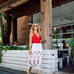 Woman in a red top and white skirt with red patterns walking outside a rustic building.