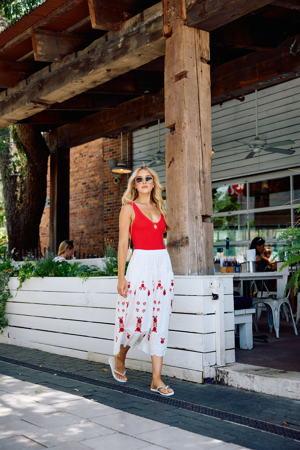 Woman in a red top and white skirt with red patterns walking outside a rustic building.