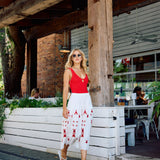 Woman in a red top and white skirt with red patterns walking outside a rustic building.