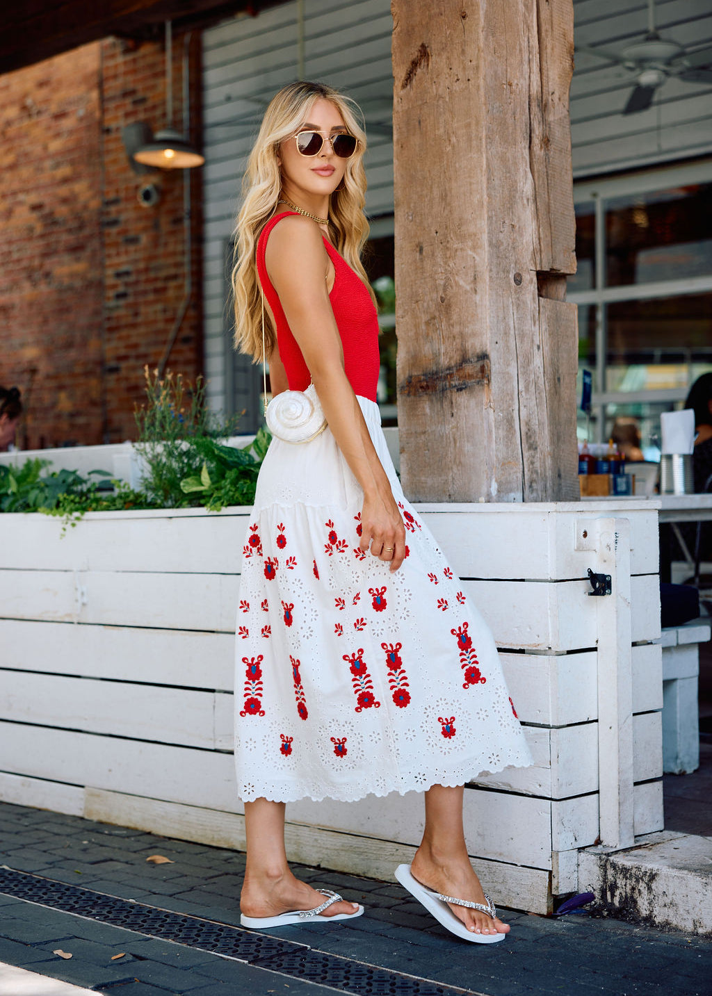Woman in a red top and white dress with red patterns with silver and white flip flops standing outdoors.