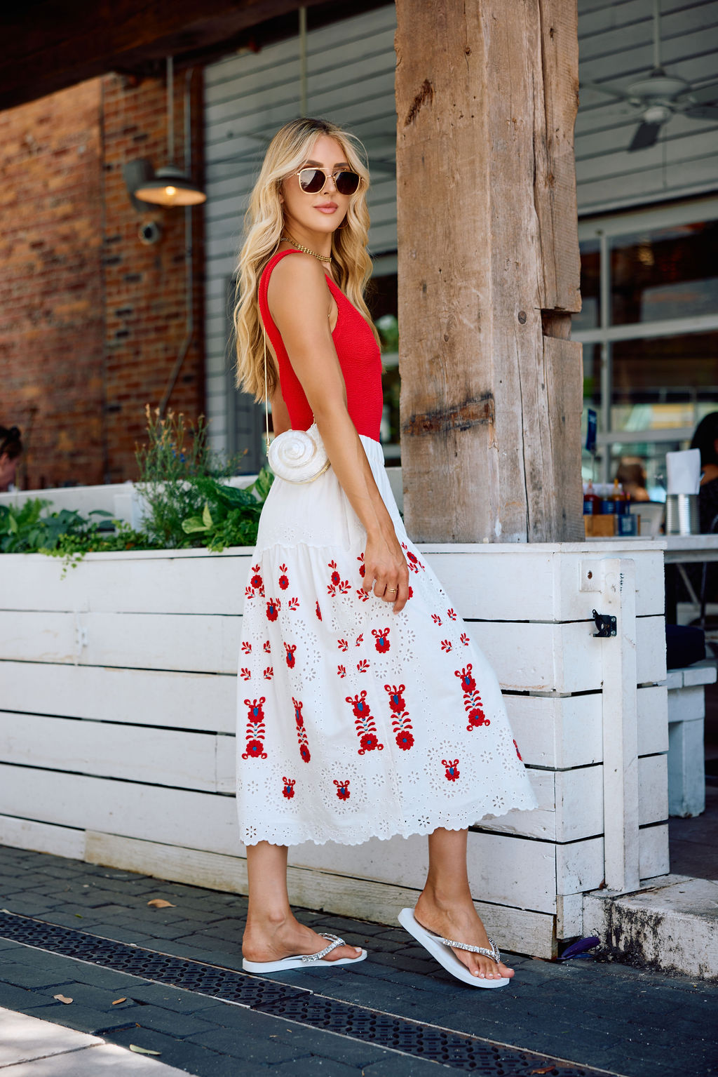 Woman in a red top and white dress with red patterns with silver and white flip flops standing outdoors.