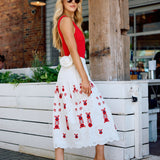 Woman in a red top and white dress with red patterns with silver and white flip flops standing outdoors.