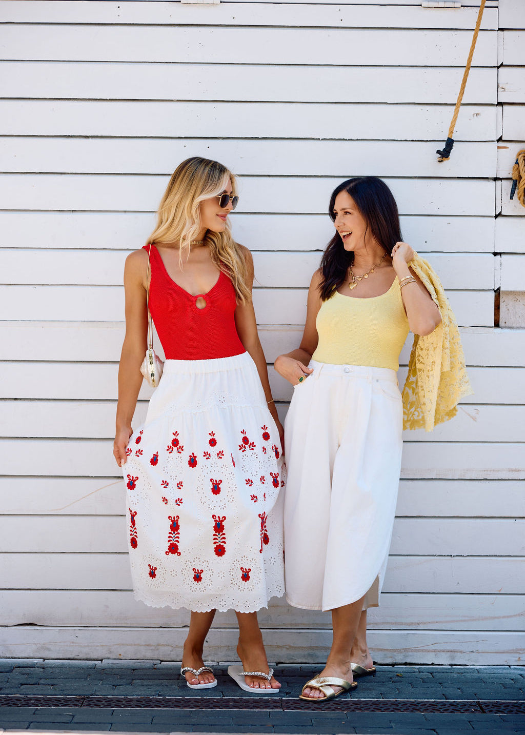 Two women standing on a wooden deck, wearing colorful sets against a white wall.