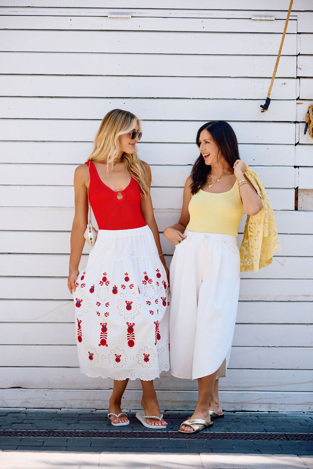 Two women standing on a wooden deck, wearing colorful sets against a white wall.