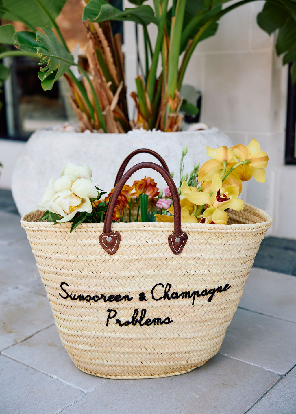 Woven basket with flowers and text 'Sunscreen & Champagne Problems' on a tiled floor.