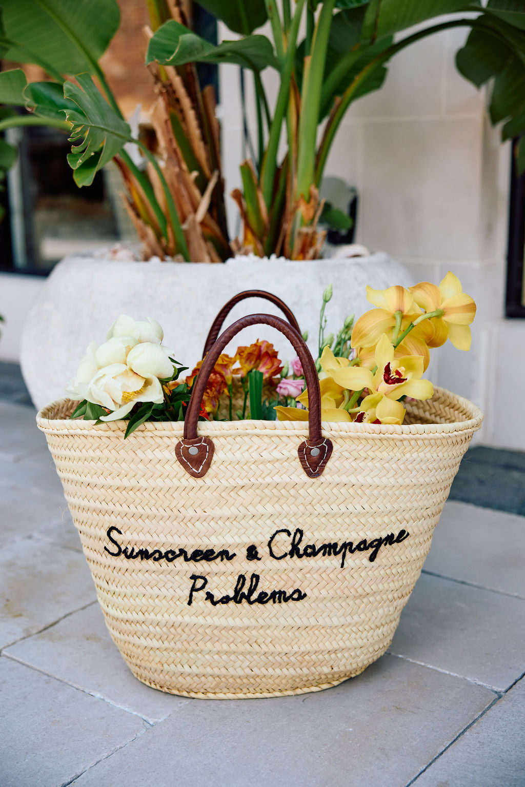 Woven basket with flowers and text 'Sunscreen & Champagne Problems' on a tiled floor.