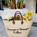Woven basket with flowers and text 'Sunscreen & Champagne Problems' on a tiled floor.
