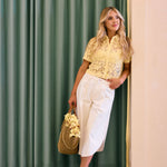 Woman in a yellow blouse and white pants standing in front of a green curtain.
