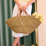 Person holding a woven handbag with a floral arrangement, wearing a yellow top , gold floral heels and white pants.