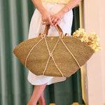 Person holding a woven handbag with a floral arrangement, wearing a yellow top, gold shoes and white pants.