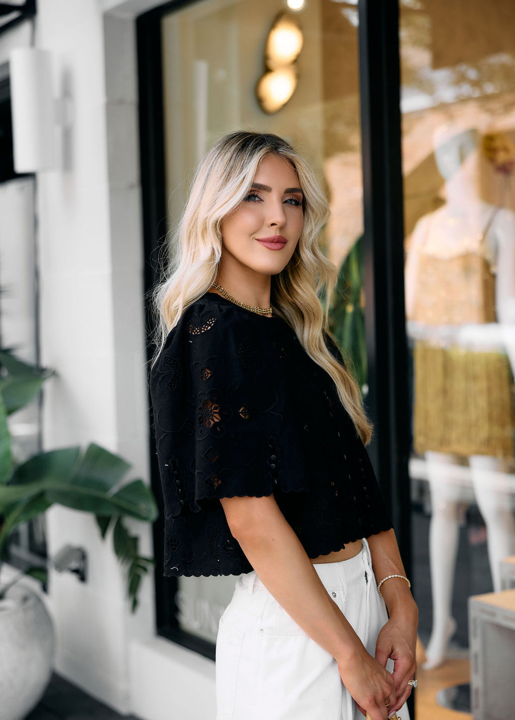 Woman in an embroidered black top and white pants standing in front of a store window.