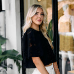 Woman in an embroidered black top and white pants standing in front of a store window.