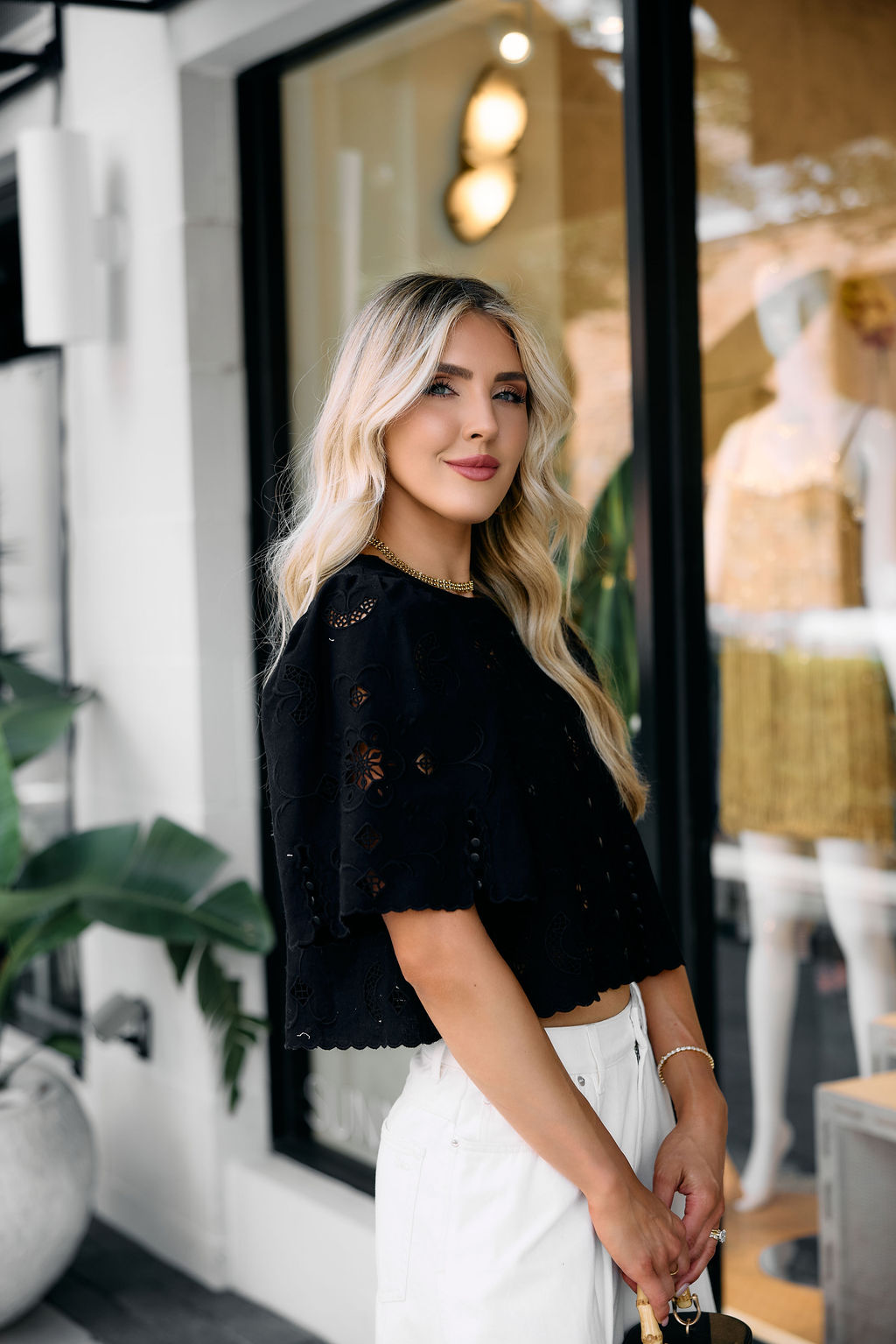 Woman in an embroidered black top and white pants standing in front of a store window.