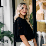 Woman in an embroidered black top and white pants standing in front of a store window.