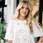 Woman wearing a white lace top with floral patterns in an outdoor setting.