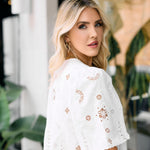 Woman wearing a white lace top with floral patterns in an outdoor setting.