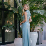 Woman in a light blue maxi skirt and beaded top standing among large plants outdoors.
