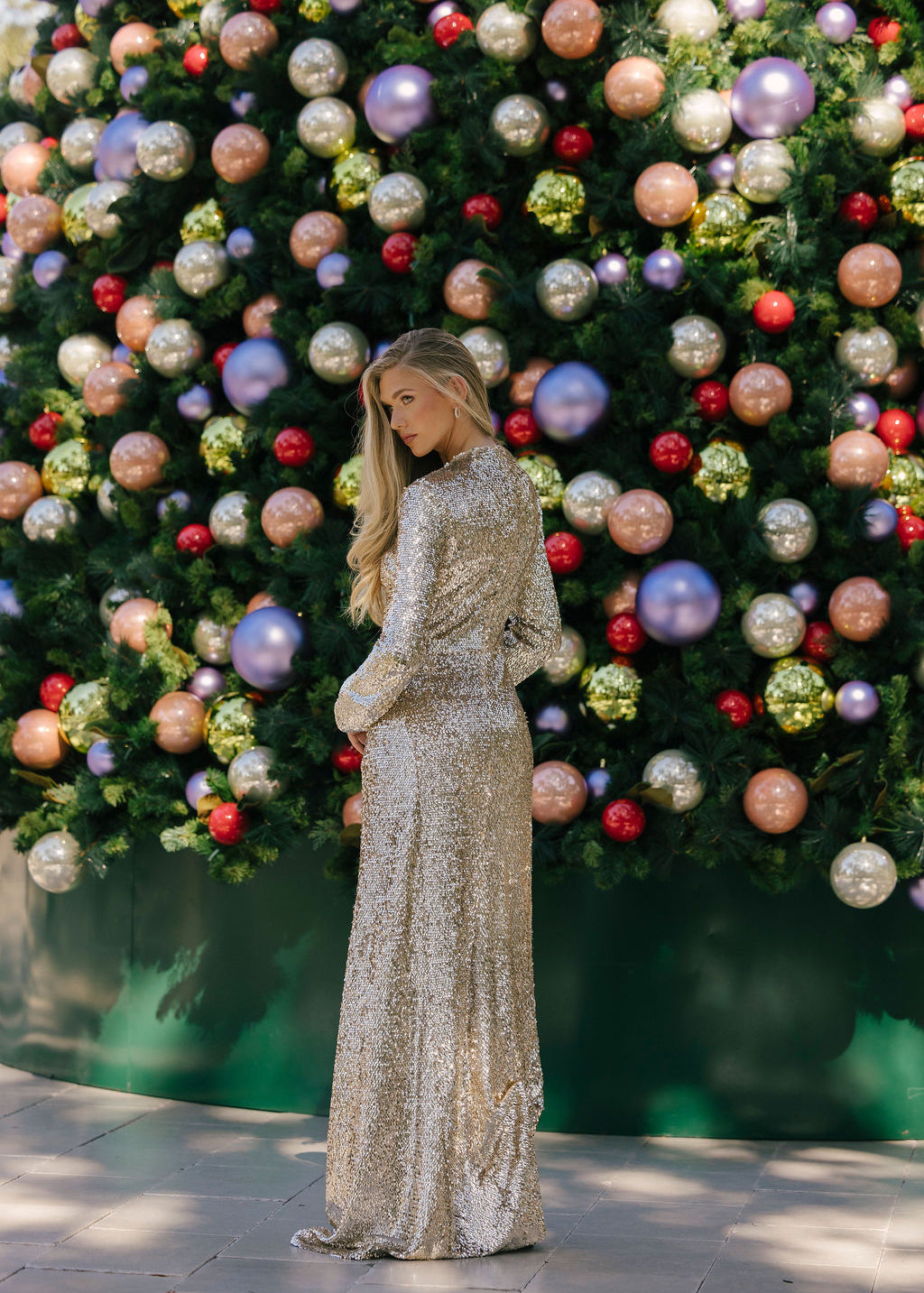 Woman in a sparkling silver dress standing in front of a decorated Christmas tree with colorful ornaments.