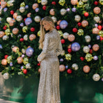 Woman in a sparkling silver dress standing in front of a decorated Christmas tree with colorful ornaments.