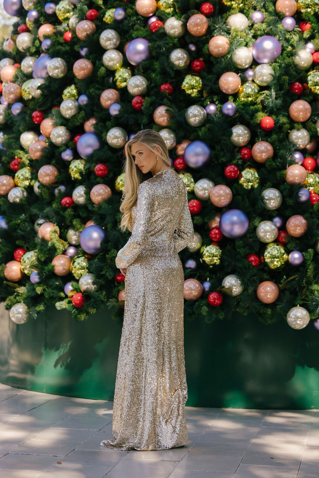 Woman in a sparkling silver dress standing in front of a decorated Christmas tree with colorful ornaments.