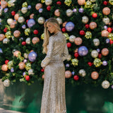 Woman in a sparkling silver dress standing in front of a decorated Christmas tree with colorful ornaments.