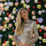 Woman in a sparkling silver dress standing in front of a decorated Christmas tree with colorful ornaments.