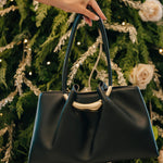 Black handbag held by a hand in front of a decorated Christmas tree.