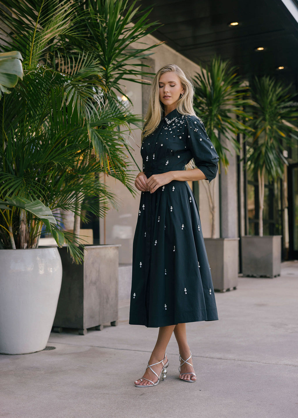 Woman in a black dress with white patterns standing outdoors among potted plants.