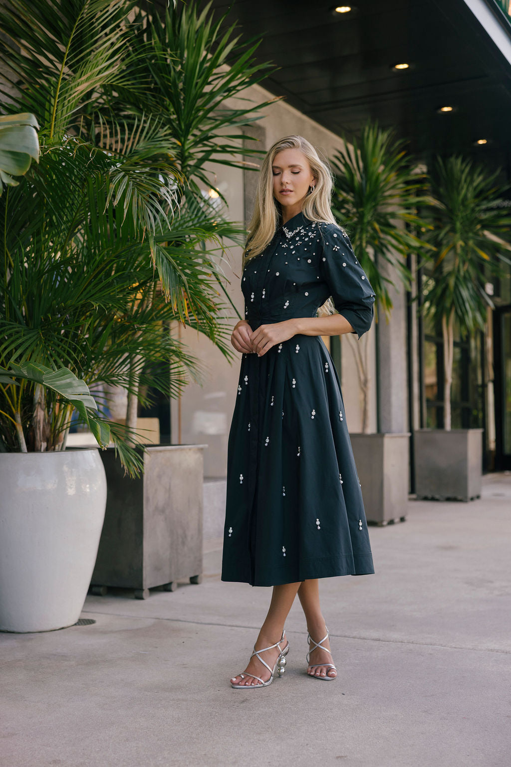 Woman in a black dress with white patterns standing outdoors among potted plants.