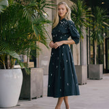 Woman in a black dress with white patterns standing outdoors among potted plants.