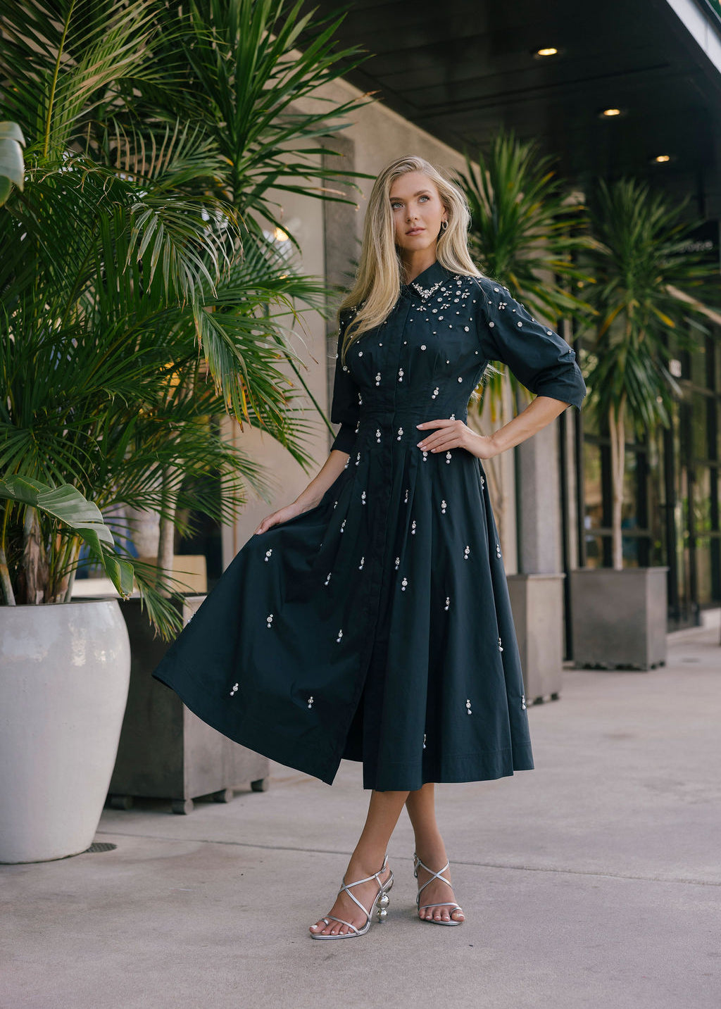 Woman in a black embellished dress standing outdoors with plants and a building in the background