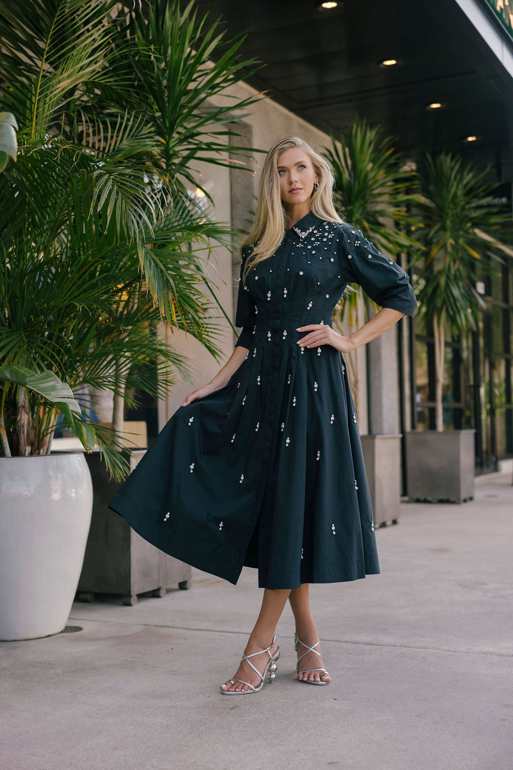 Woman in a black embellished dress standing outdoors with plants and a building in the background