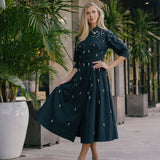 Woman in a black embellished dress standing outdoors with plants and a building in the background