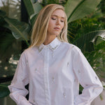 Woman wearing a white embellished shirt with green foliage in the background