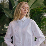 Woman wearing a white embellished shirt with green foliage in the background