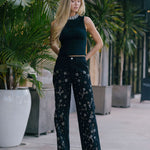 Woman in a black outfit with floral patterns  embellished and silver heels standing outdoors among plants.