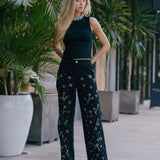 Woman in a black outfit with floral patterns embellished standing outdoors among plants.