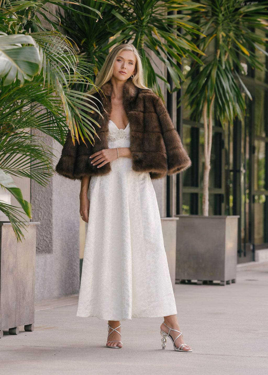 Woman in a white dress with a fur coat and silver heels standing in an outdoor setting with plants.
