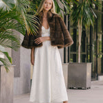 Woman in a white dress with a fur coat and silver heels standing in an outdoor setting with plants.